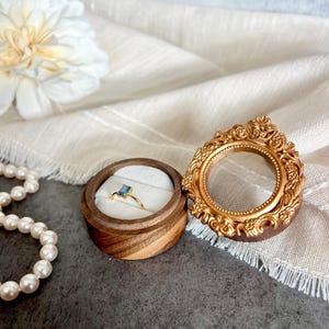 May include: A gold ring with a rectangular blue gemstone sits in a wooden ring box with a white interior. A decorative gold frame and a strand of pearls are also visible. The background includes a white flower and a cream-colored cloth.