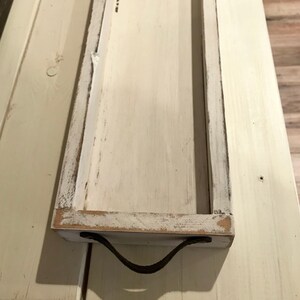 May include: White wooden tray with a distressed finish and a brown leather handle. The tray is rectangular and has a raised edge.