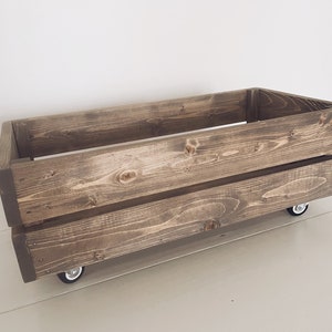 May include: A rectangular wooden crate with a dark brown stain, mounted on four small caster wheels. The crate has a simple, open design, with visible wood grain and knots. The crate is designed for storage.