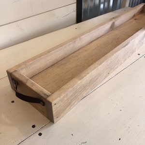 May include: A long, rectangular wooden tray with a light brown stain. The tray has a leather handle on one side and is perfect for serving or displaying items.