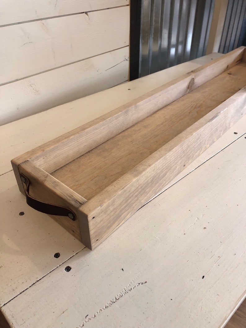 May include: A long, rectangular wooden tray with a light brown stain. The tray has a leather handle on one side and is perfect for serving or displaying items.