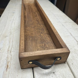 May include: A long, rectangular wooden tray with a dark brown stain. The tray has a black leather handle with brass rivets. The tray is sitting on a white wooden surface.