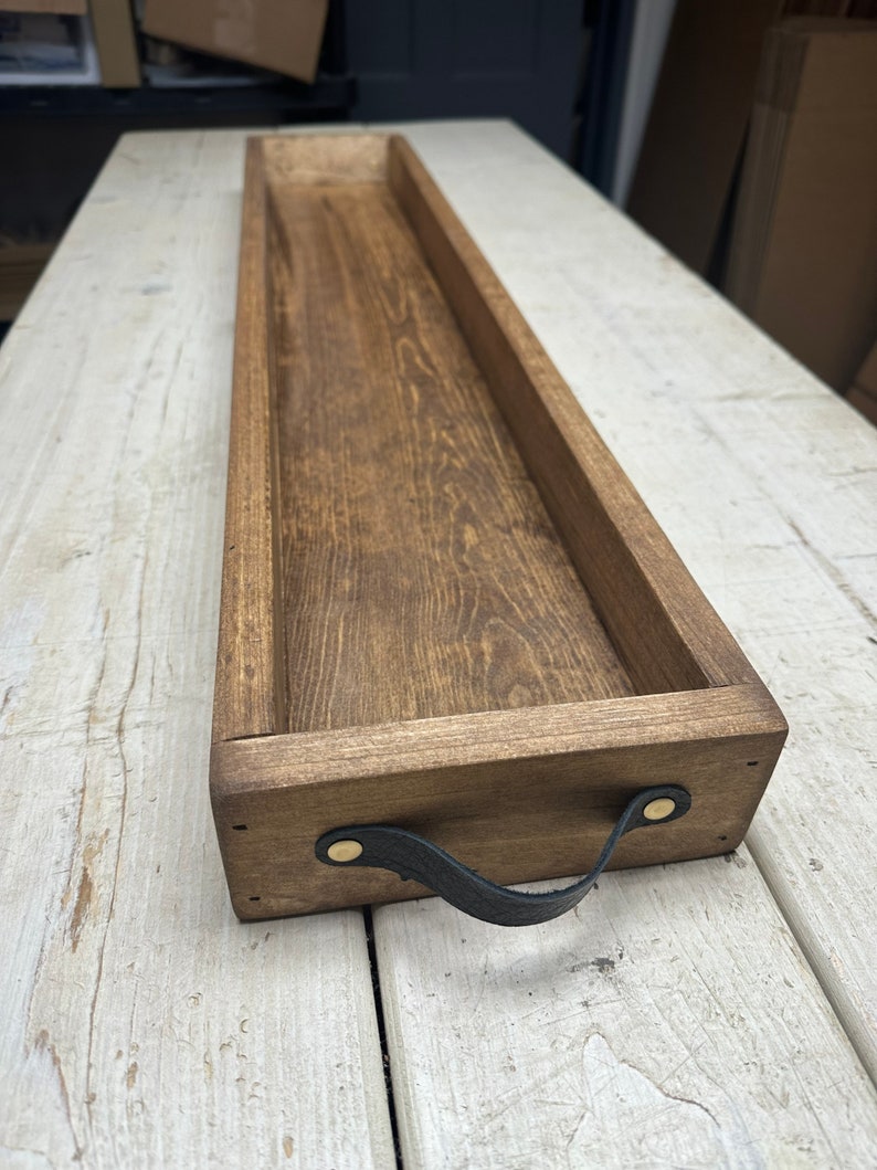 May include: A long, rectangular wooden tray with a dark brown stain. The tray has a black leather handle with brass rivets. The tray is sitting on a white wooden surface.