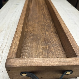 May include: A long, rectangular wooden tray with a dark brown stain. The tray has a leather handle with brass accents on each side.
