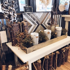 May include: A white wooden table with a wooden tray filled with white ceramic containers and greenery. The table is decorated with wicker baskets, a sign that reads "Table $145", and a wooden sign with the text "Autumn the Year's last Sweetest Smile".