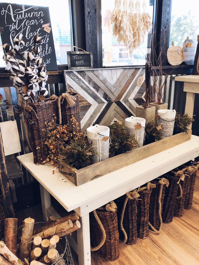 May include: A white wooden table with a wooden tray filled with white ceramic containers and greenery. The table is decorated with wicker baskets, a sign that reads "Table $145", and a wooden sign with the text "Autumn the Year's last Sweetest Smile".