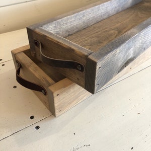 May include: Two wooden trays with leather handles. The trays are stacked on top of each other. The trays are made of reclaimed wood and have a rustic finish.