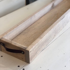 May include: A long, rectangular wooden tray with a light brown stain. The tray has a leather handle on one end and is made of unfinished wood.