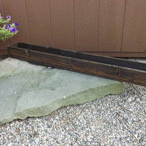 May include: A long, dark brown wooden planter box with a rustic finish. The planter is sitting on a large, flat rock.