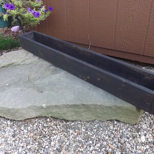 May include: A long, black wooden planter box with a rustic finish. The planter is sitting on a large, flat rock.