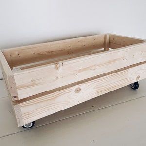 May include: A natural wood storage crate with wheels. The crate is made of unfinished wood and has a rectangular shape. It has two small wheels on the bottom.