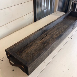 May include: A long, dark brown wooden tray with leather handles. The tray has a rustic, farmhouse style.