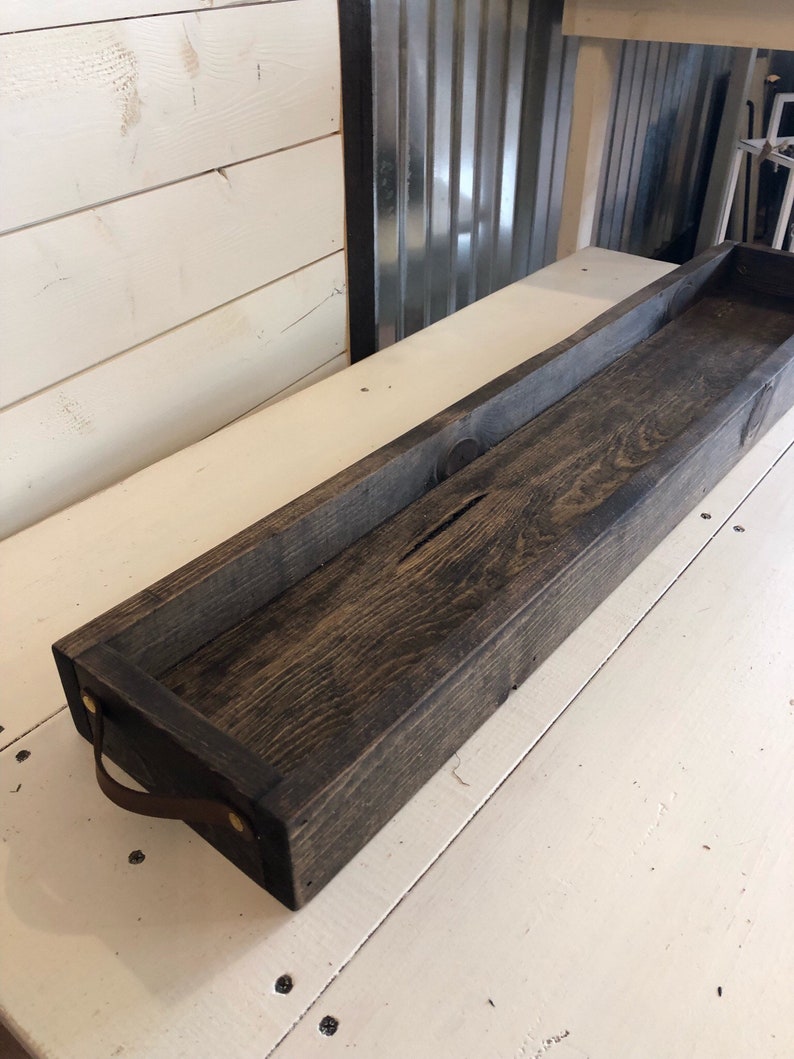 May include: A long, dark brown wooden tray with leather handles. The tray has a rustic, farmhouse style.