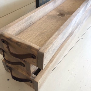 May include: Two wooden trays with leather handles. The trays are stacked on top of each other. The top tray is a light brown color and the bottom tray is a darker brown color. The trays have a rustic look and are perfect for organizing and displaying items.