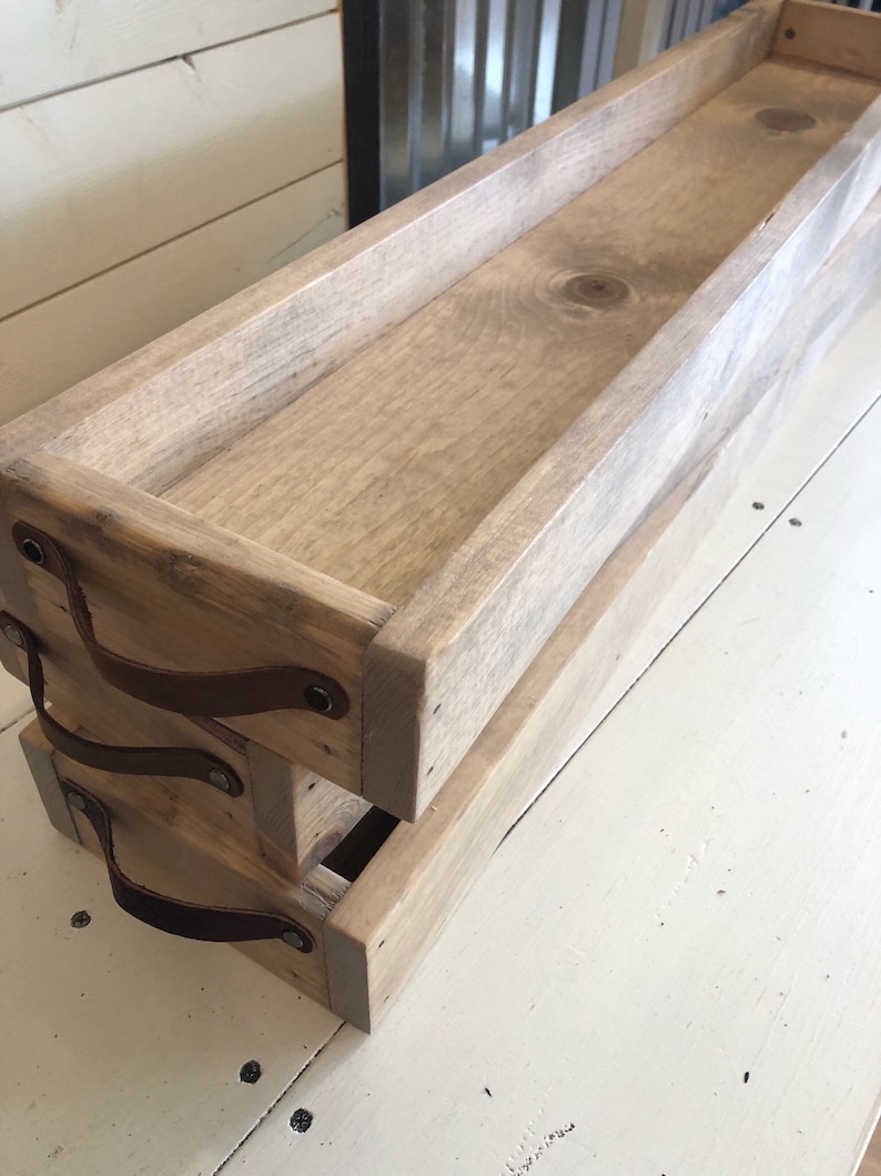 May include: Two wooden trays with leather handles. The trays are stacked on top of each other. The top tray is a light brown color and the bottom tray is a darker brown color. The trays have a rustic look and are perfect for organizing and displaying items.