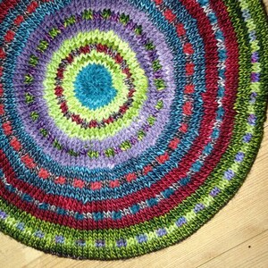 May include: A circular knitted rug with a multi-colored design. The rug has a central circle of blue surrounded by concentric circles of green, red, purple, and blue. The outer edge of the rug is green.