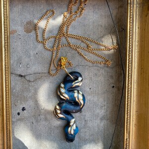 May include: A gold chain necklace with a black and blue snake pendant. The snake is decorated with gold accents and white details.