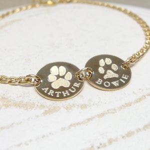 May include: A gold-toned chain bracelet featuring two circular charms. Each charm is engraved with a paw print and a name, specifically the names Arthur and Bowie, centred below the paw print designs on the polished metal surfaces.