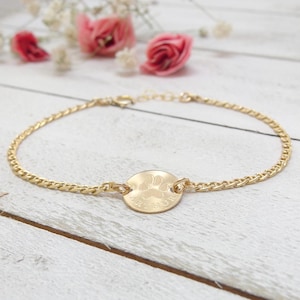 May include: A gold-coloured bracelet with a chain design. The bracelet features a circular charm with a paw print design. The background includes pink roses and white flowers on a white wooden surface.