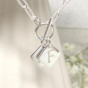 Puede incluir: Collar de plata con cierre de palanca y dos dijes cuadrados con la letra 'F' grabada.