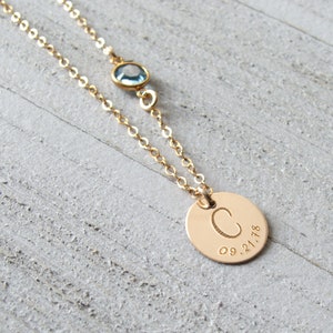 May include: Gold necklace with a small blue gemstone and a round pendant with the letter "C" and the date "09.21.18" engraved on it.