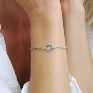May include: A silver bracelet featuring a small, round charm with a paw print design. The bracelet is delicate and sits on the wrist. The charm is centered on a fine chain.