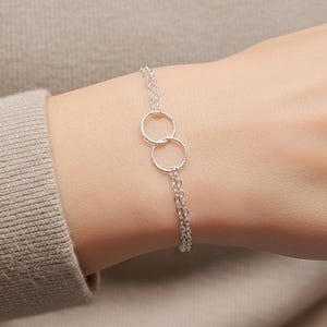 May include: A silver bracelet featuring two interlocking circles connected by a delicate chain. The bracelet is displayed on a person's wrist, with a neutral-toned sweater in the background. The jewelry is simple and elegant.