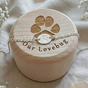 May include: A silver bracelet with a paw print charm is displayed on a wooden block engraved with a paw print and the words "Our Lovebug". The bracelet is on a light-colored fabric, with white flowers and petals scattered around.