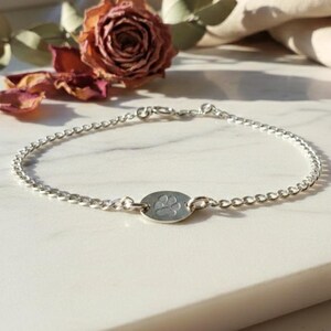 May include: A delicate silver bracelet featuring a chain design with a small, oval charm. The charm is engraved with a paw print. The bracelet is displayed on a white surface, with a dried rose and greenery in the background.