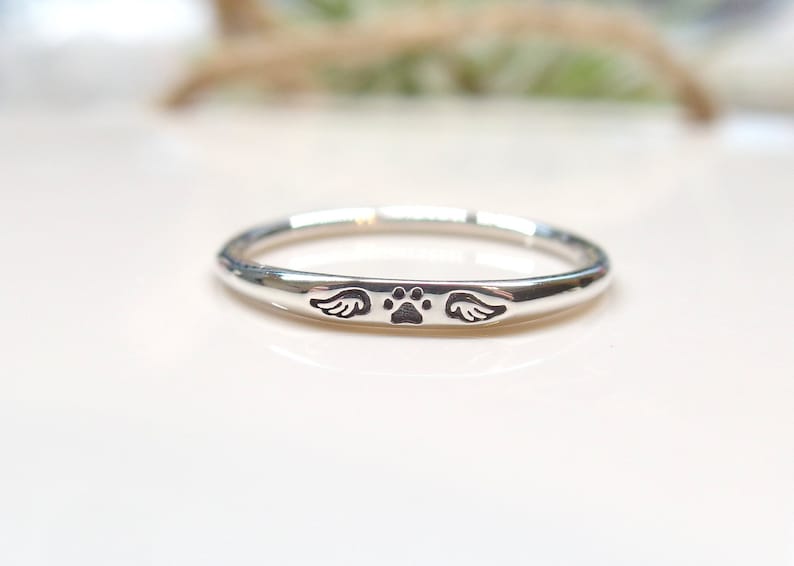 May include: A silver ring with a paw print and wings engraved on it. The ring is a simple band with a smooth finish.