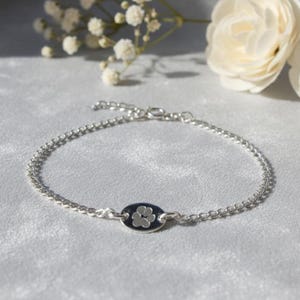 May include: A silver bracelet with a small, oval charm displaying a white paw print on a dark blue background. The bracelet is made of small, interconnected beads. The bracelet is displayed on a soft, light-coloured surface.