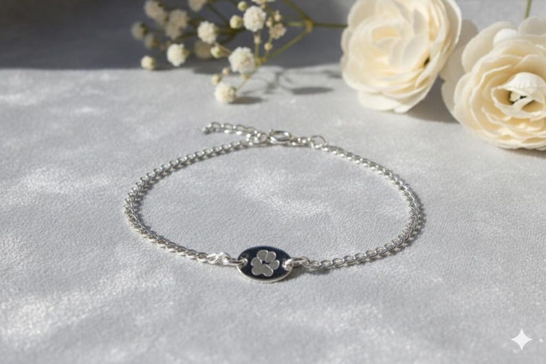 May include: A silver bracelet with a small, oval charm displaying a white paw print on a dark blue background. The bracelet is made of small, interconnected beads. The bracelet is displayed on a soft, light-coloured surface.