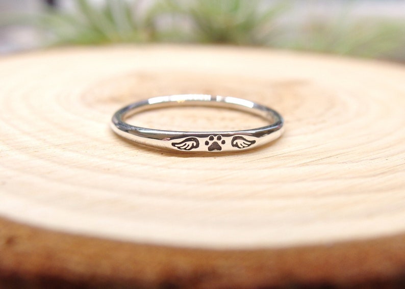 May include: A silver ring with a paw print and wings engraved on it. The ring is on a wooden surface.