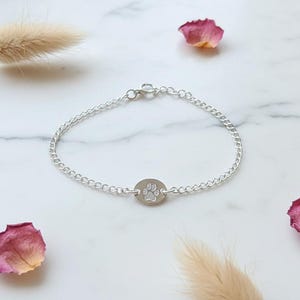 May include: A silver chain bracelet with a circular charm featuring a paw print. The bracelet is displayed on a white marble surface, decorated with dried rose petals and a sprig of dried grass.