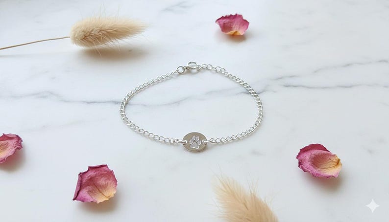 May include: A silver chain bracelet with a circular charm featuring a paw print. The bracelet is displayed on a white marble surface, decorated with dried rose petals and a sprig of dried grass.