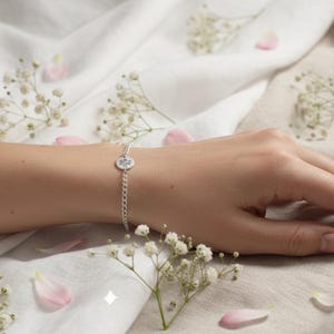 May include: A silver bracelet with a small, round charm is shown on a wrist. The bracelet is on a chain and the charm has a design. The background is a white and beige fabric with scattered pink petals and small white flowers.