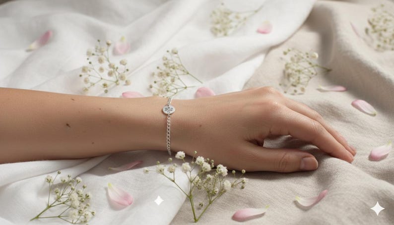 May include: A silver bracelet with a small, round charm is shown on a wrist. The bracelet is on a chain and the charm has a design. The background is a white and beige fabric with scattered pink petals and small white flowers.