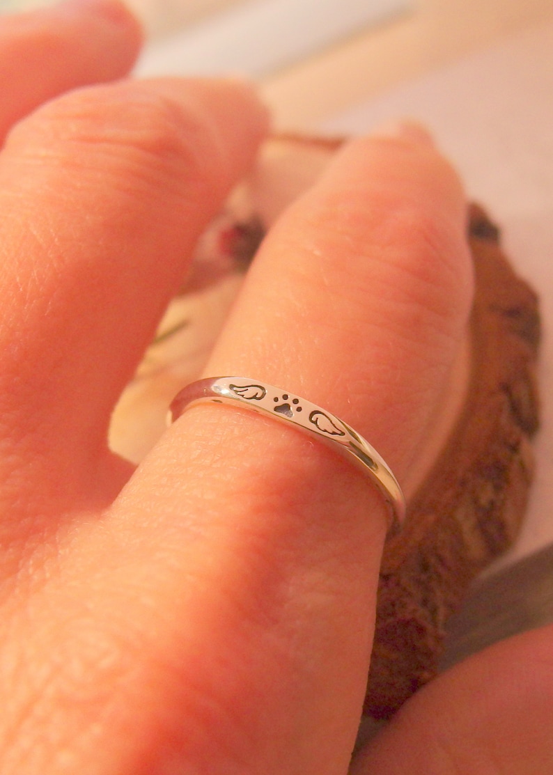 May include: A silver ring with a paw print and two wings engraved on it. The ring is worn on a finger.