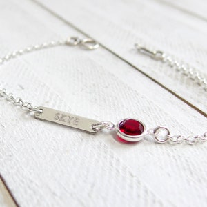 May include: A silver chain bracelet with a small red gemstone and a personalized bar charm engraved with the name "SKYE".