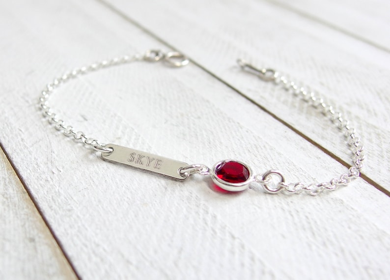 May include: A silver chain bracelet with a small red gemstone and a personalized bar charm engraved with the name "SKYE".
