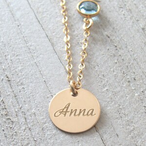 May include: Gold necklace with a round pendant engraved with the name "Anna" and a small light blue gemstone.