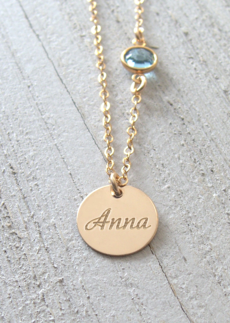 May include: Gold necklace with a round pendant engraved with the name "Anna" and a small light blue gemstone.