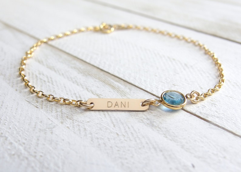 May include: Gold chain bracelet with a small blue gemstone and a personalized bar charm engraved with the name "DANI".