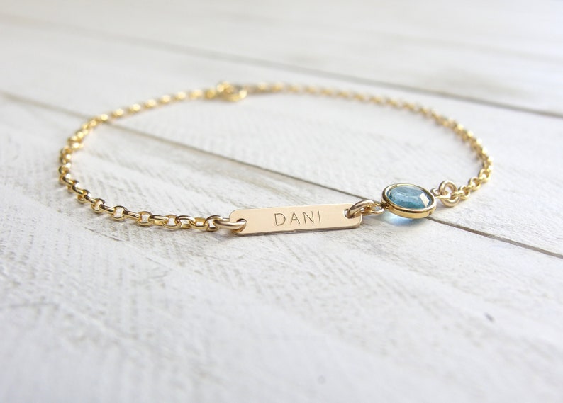 May include: A gold chain bracelet with a small gold bar charm engraved with the name "DANI" and a light blue gemstone charm.