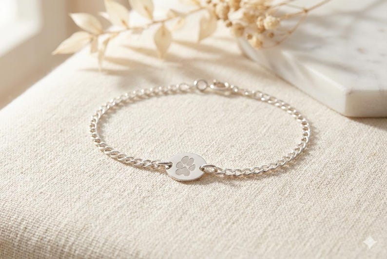 May include: A silver bracelet with a delicate chain and a circular charm featuring a paw print. The bracelet is displayed on a neutral-toned surface, with soft lighting and a blurred background.