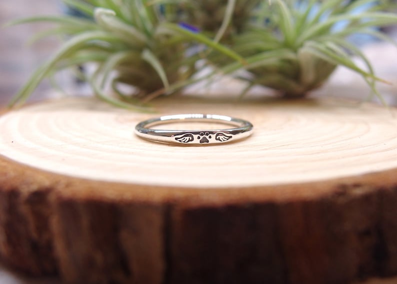 May include: A silver ring with a paw print and wings engraved on it. The ring is on a wooden surface.