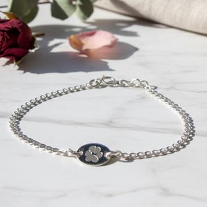 May include: Silver chain bracelet with an oval charm featuring a paw print design. The bracelet is displayed on a white marble surface, with a dark red rose and petals in the background.