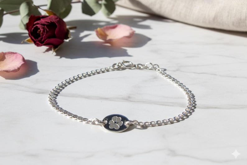 May include: Silver chain bracelet with an oval charm featuring a paw print design. The bracelet is displayed on a white marble surface, with a dark red rose and petals in the background.