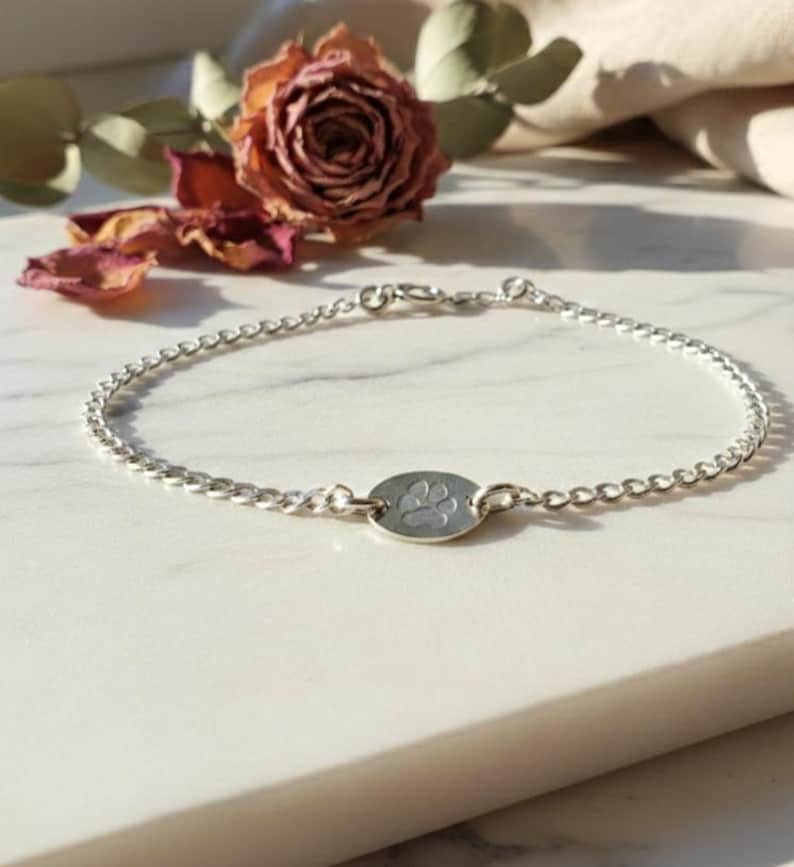 May include: A delicate silver bracelet featuring a chain design with a small, oval charm. The charm is engraved with a paw print. The bracelet is displayed on a white surface, with a dried rose and greenery in the background.