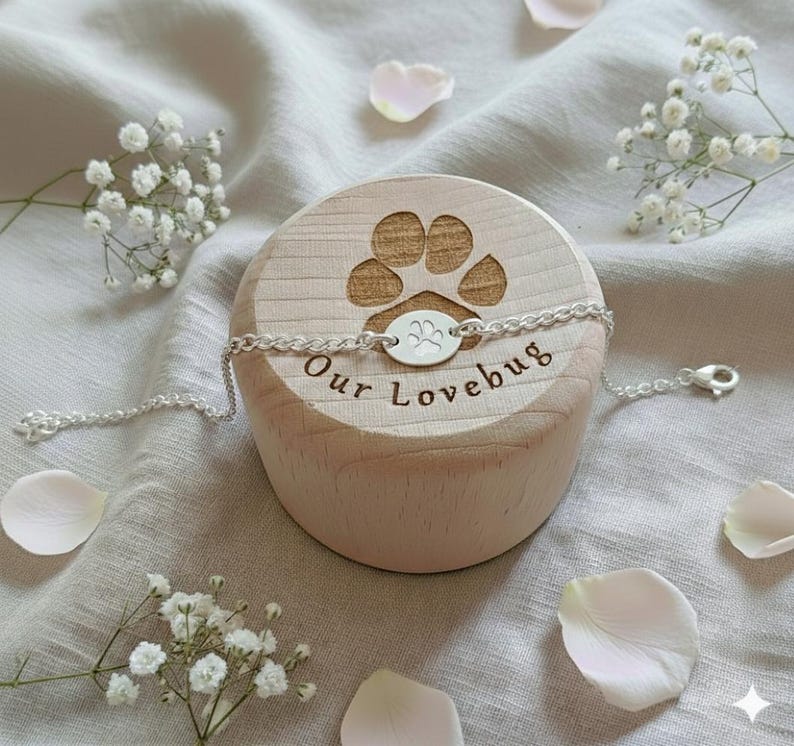 May include: A silver bracelet with a paw print charm is displayed on a wooden block engraved with a paw print and the words "Our Lovebug". The bracelet is on a light-colored fabric, with white flowers and petals scattered around.